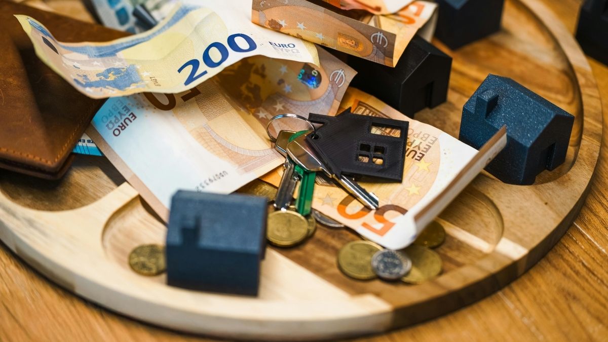 Chiavi con portachiavi a forma di casa, banconote in euro, monete e piccoli modelli di abitazioni su un vassoio di legno.