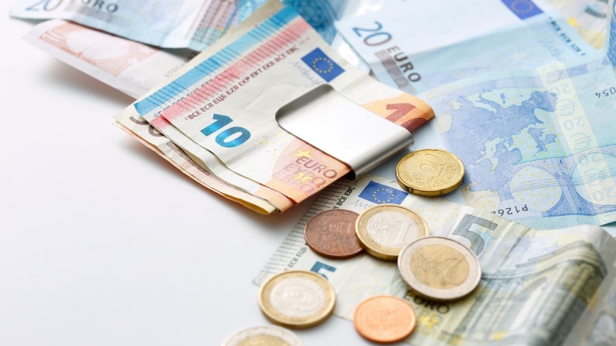 Banconote in euro trattenute da un fermasoldi metallico e monete sparse su altre banconote, su fondo chiaro.