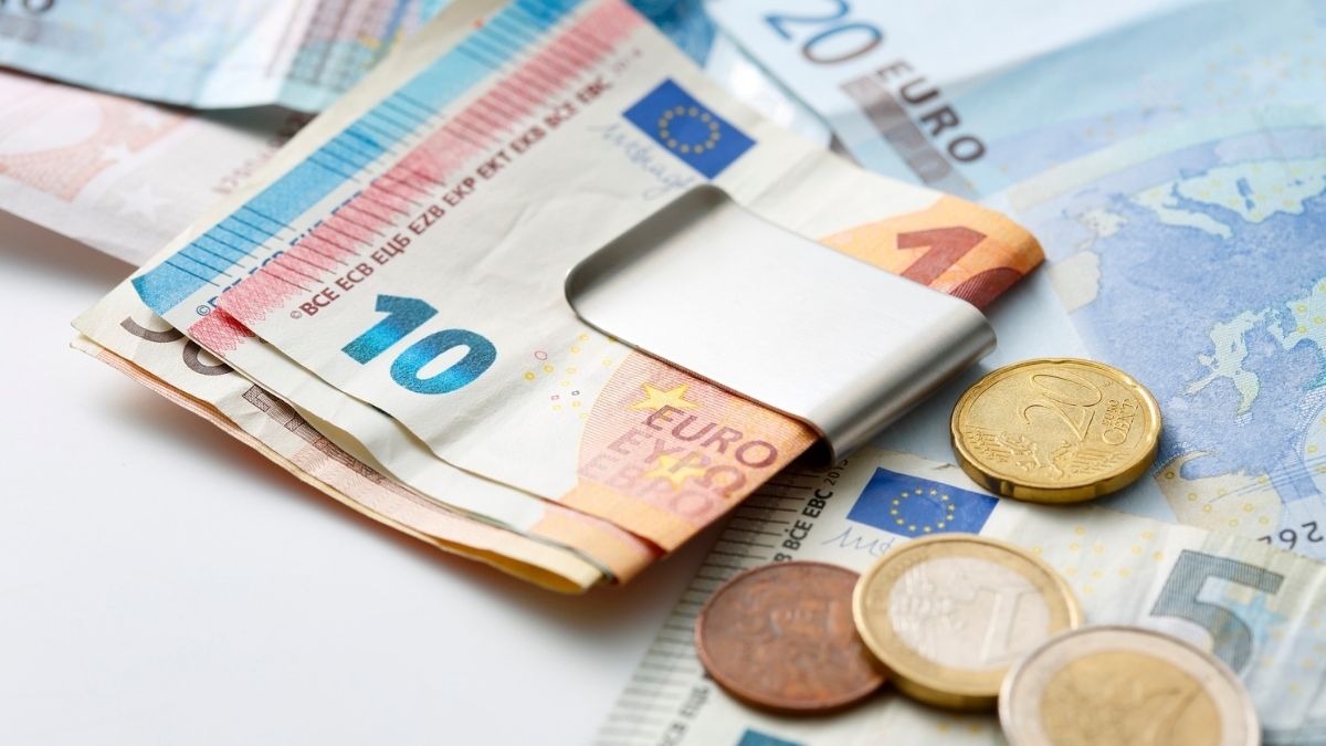 Dettaglio di banconote in euro piegate in un fermasoldi argentato, accanto a monete su fondo bianco.