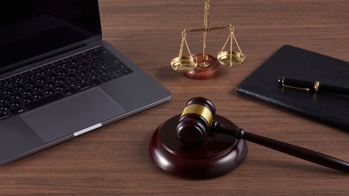 Martelletto su una base, bilancia dorata, computer portatile e penna su un quaderno nero, disposti su una scrivania di legno.