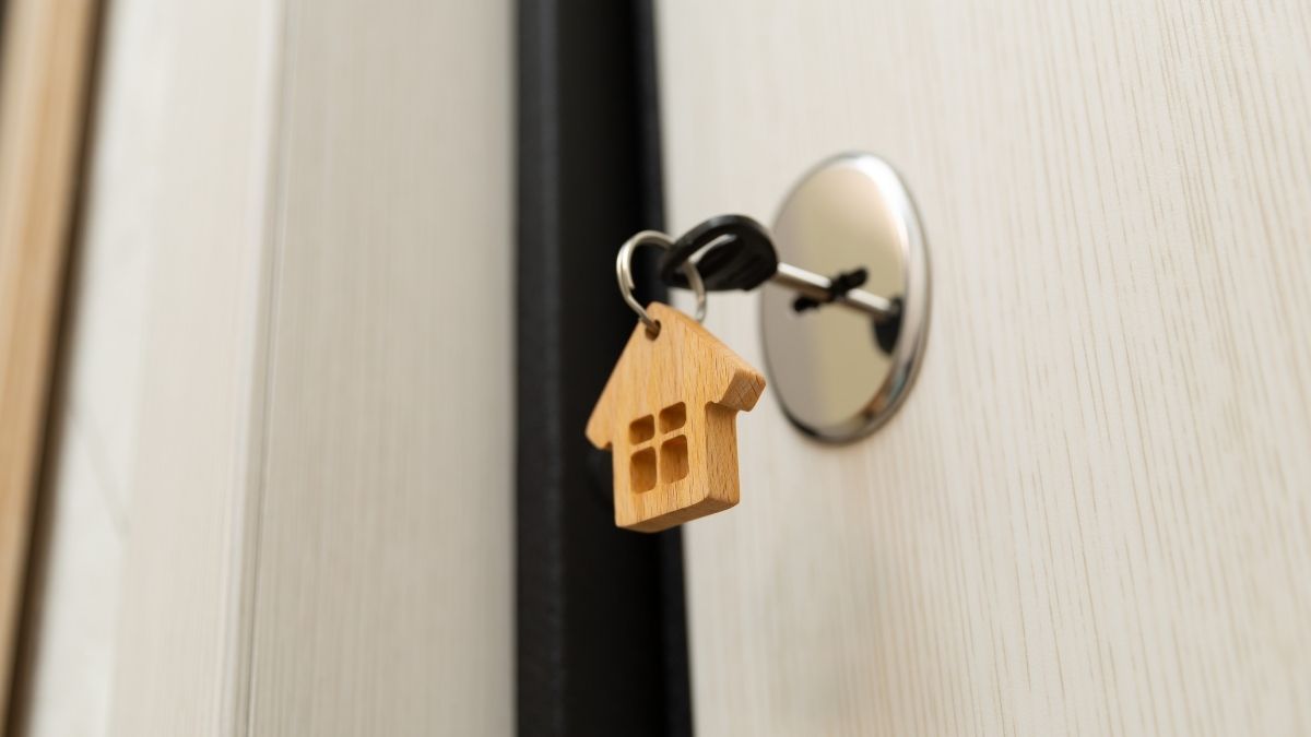 Chiave con portachiavi di legno a forma di casa inserita nella serratura di una porta chiara socchiusa.