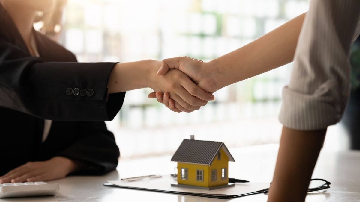 Due persone si stringono la mano sopra una scrivania con un modellino giallo di casa e documenti.