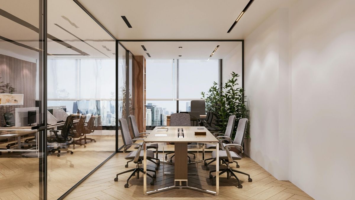 Sala riunioni luminosa con tavolo in legno, sedie grigie, piante e pareti di vetro affacciate su postazioni di lavoro.