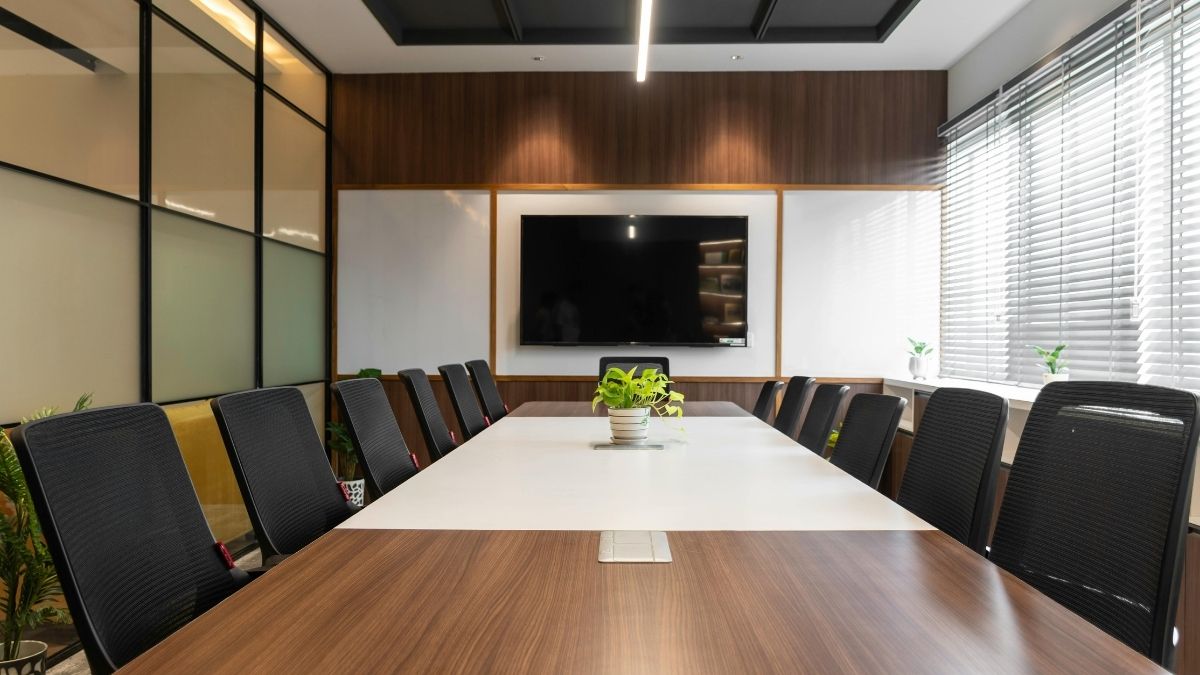 Sala riunioni vuota con lungo tavolo in legno, sedie nere, una pianta al centro e un grande schermo spento sulla parete.