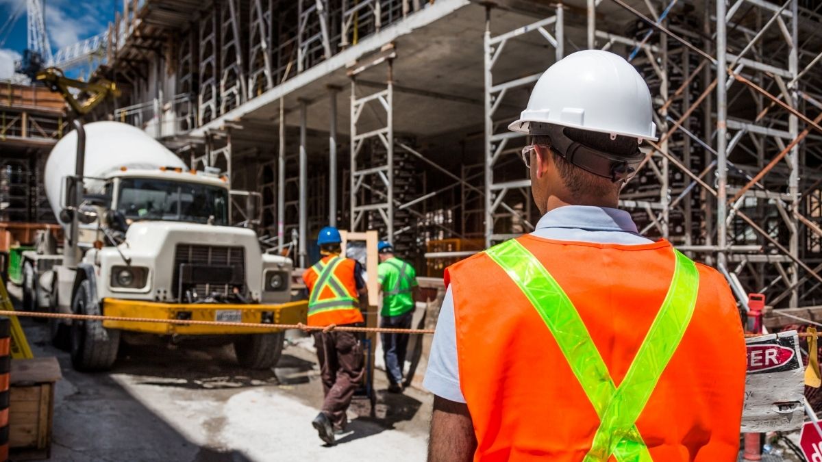 Persona di spalle con casco bianco e gilet arancione in un cantiere con ponteggi, lavoratori e un’autobetoniera.