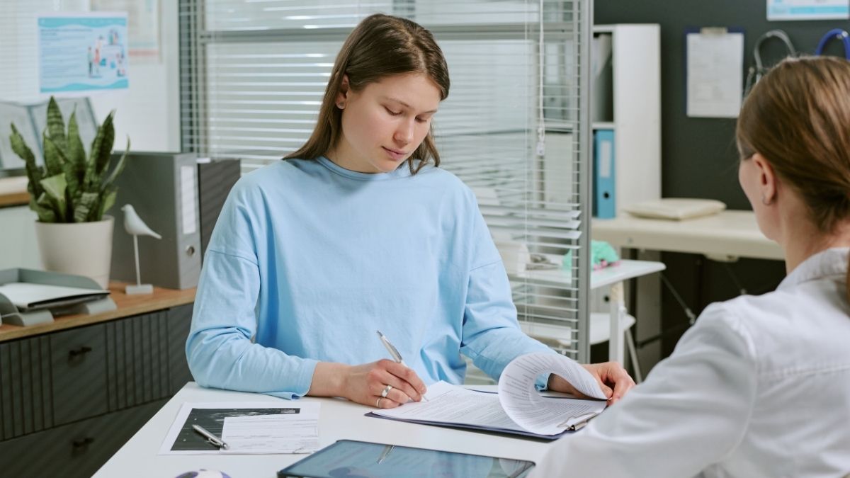 Donna in maglia azzurra scrive su documenti davanti a una persona in camice bianco, con un tablet sulla scrivania.
